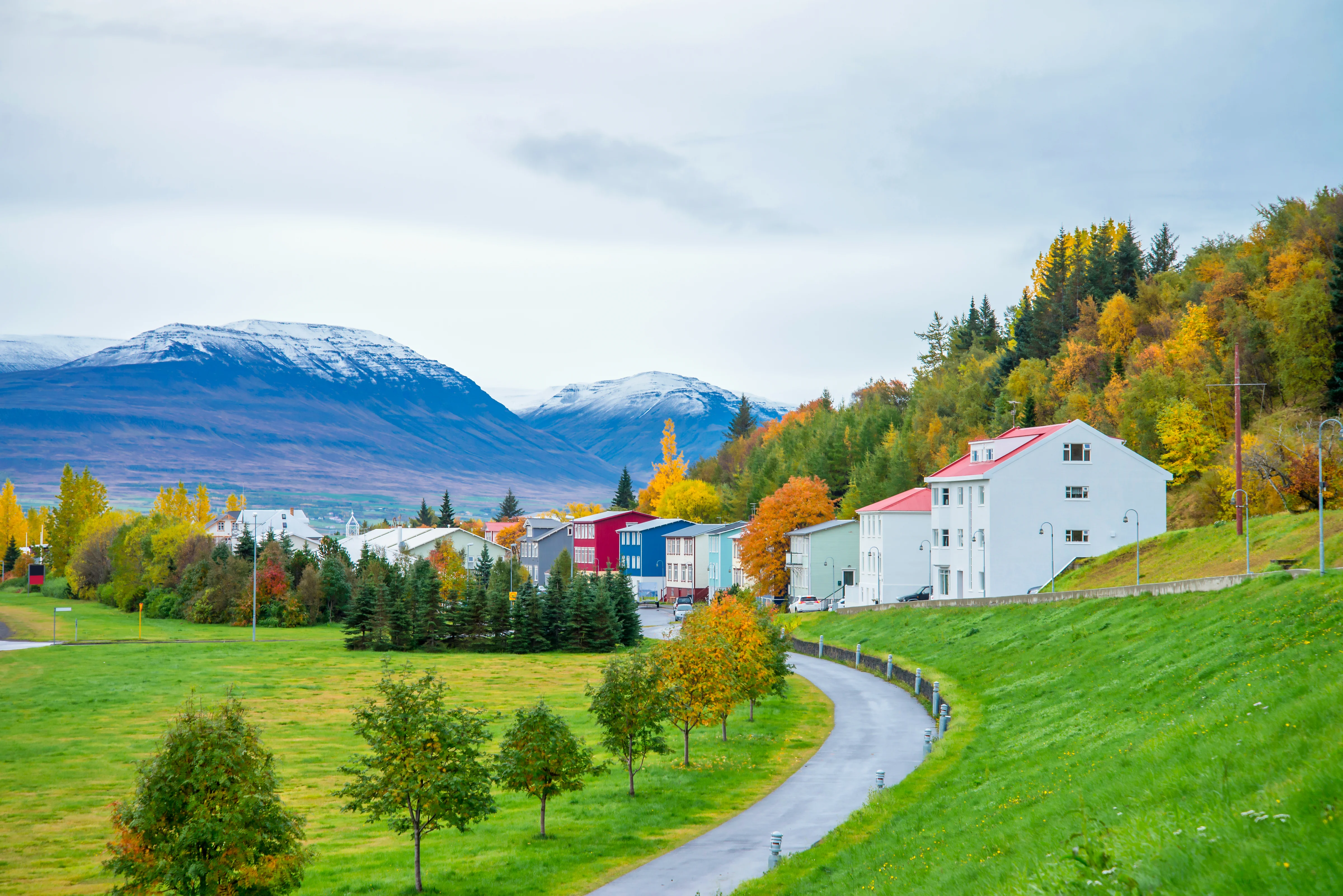 A scenic street view in Akureyri during autumn, with brightly colored houses, green fields, and snow-covered mountains in the background.