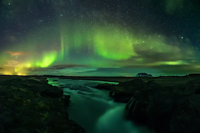 Vibrant Northern Lights reflecting over a flowing Icelandic river at night, a stunning view of Iceland's natural beauty during a scenic tour.