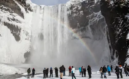 Visitors admiring the icy Skógafoss waterfall with a rainbow in the mist, during a winter tour in Iceland.