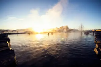 People relaxing in the warm waters of the Secret Lagoon in Flúðir, Iceland, at sunrise, surrounded by mist and a peaceful morning glow over the geothermal pool.