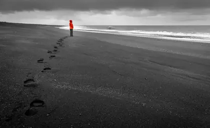 Person in bright orange jacket stands on Vik's black sand beach with footsteps behind, under stormy grey sky in Iceland.