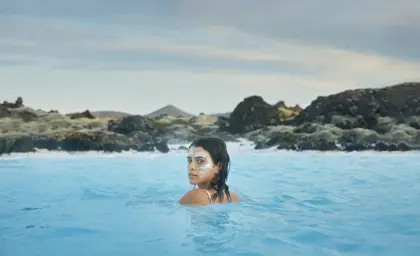 Woman with a white silica face mask relaxing in the Blue Lagoon, a geothermal spa in Iceland, surrounded by volcanic rocks and distant hills under a cloudy sky.