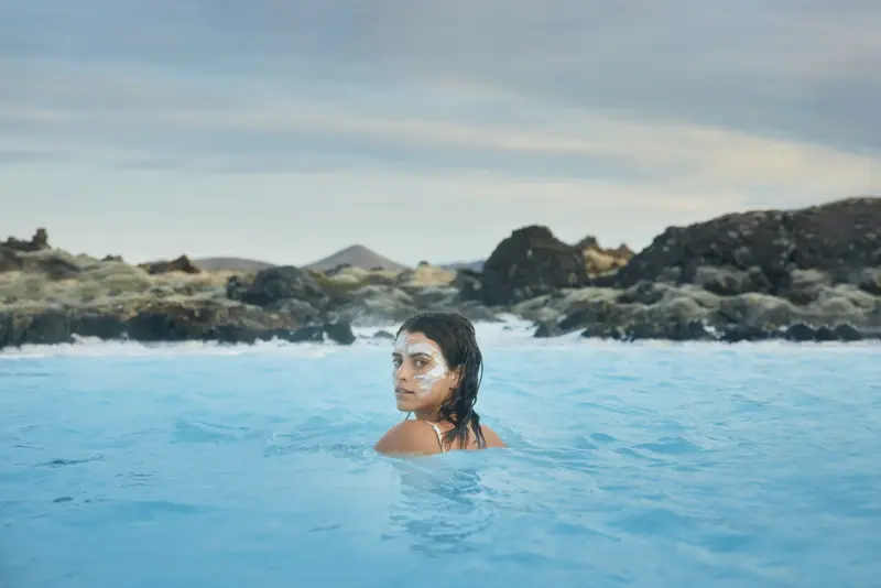 Woman with a white silica face mask relaxing in the Blue Lagoon, a geothermal spa in Iceland, surrounded by volcanic rocks and distant hills under a cloudy sky.