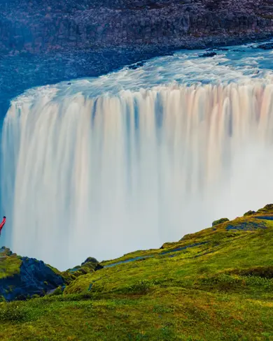 View of Dettifoss Waterfall where a hiker stands triumphantly in front of the most powerful waterfall in Europe, located in Iceland's Vatnajökull National Park, with mist rising and vibrant greenery surrounding the scene.
