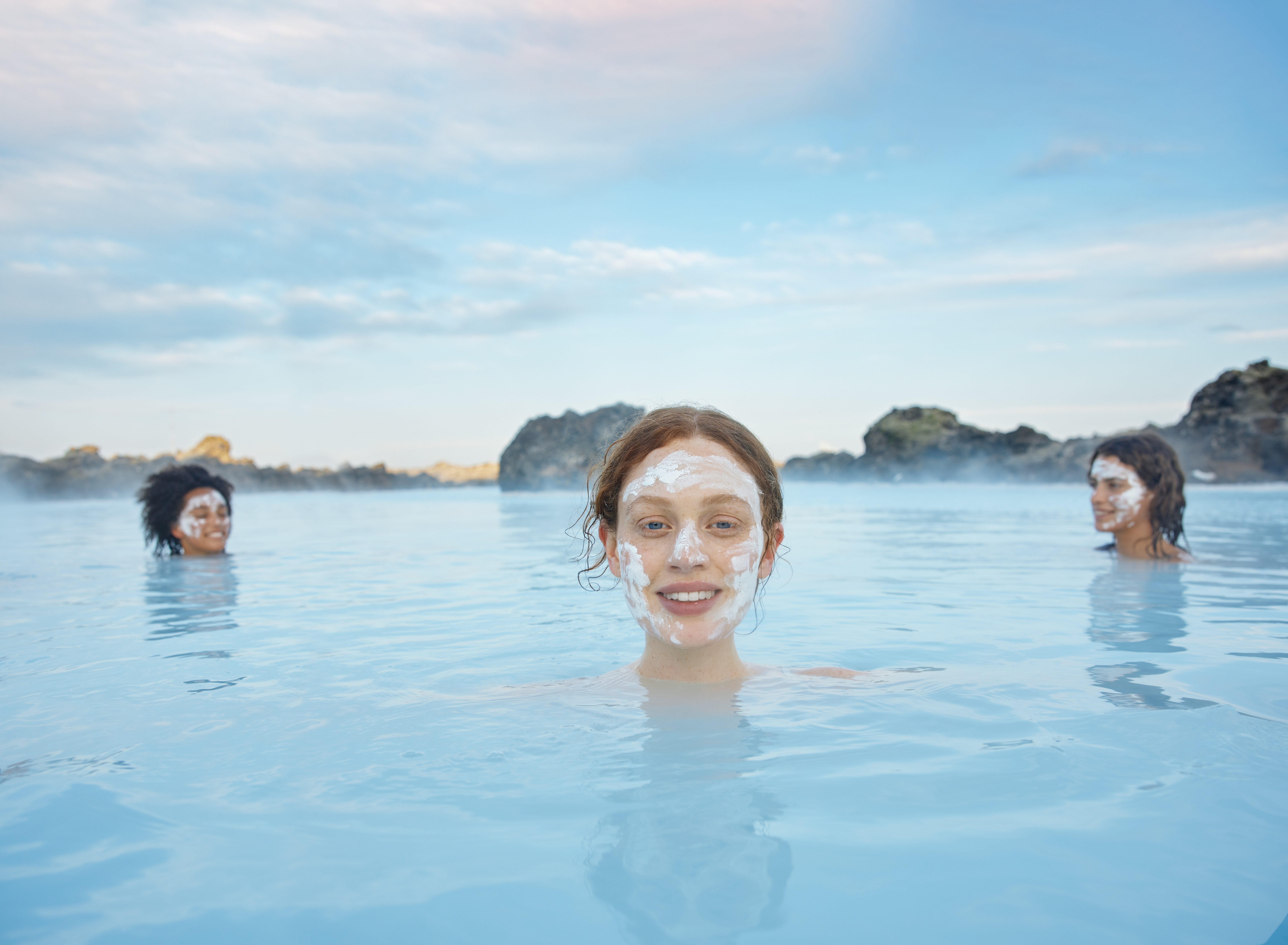 A woman with a facial mud mask enjoys the serene waters of the Blue Lagoon in Iceland, with two other guests in the background, surrounded by natural rock formations.