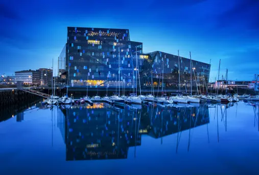 Harpa Concert Hall in Reykjavík, Iceland, at night with a view of its glass facade reflecting on the calm waters of the harbor with sailboats moored nearby.