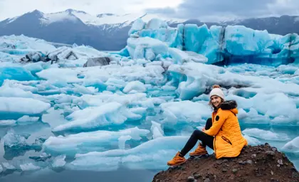 Smiling woman in a yellow parka sitting by Jokulsarlon Glacier Lagoon surrounded by floating icebergs in Iceland.