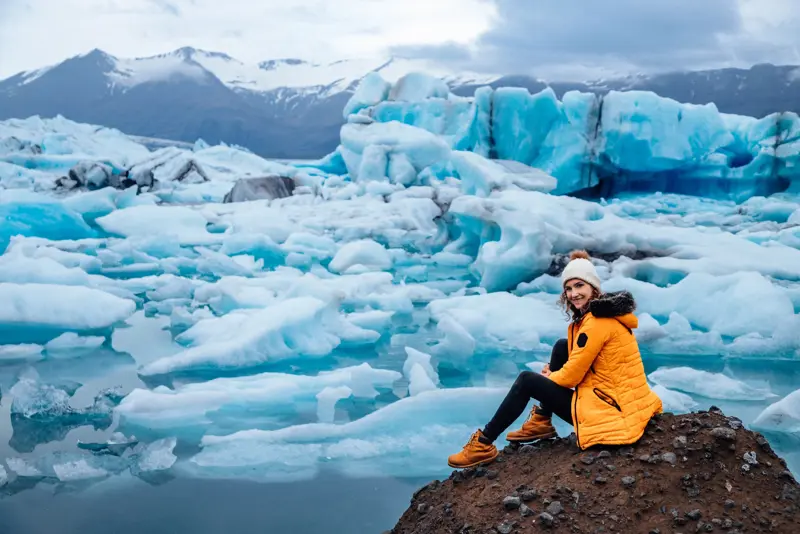 Smiling woman in a yellow parka sitting by Jokulsarlon Glacier Lagoon surrounded by floating icebergs in Iceland.