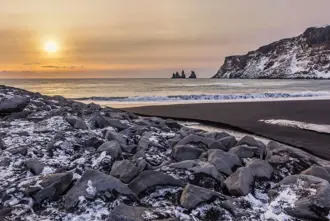 Vikurfjara black sand beach in winter with light snow covering the ground and view of Reynisdrangar at sunset.