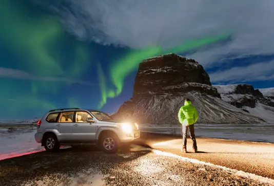 Man hunting for the northern lights in Iceland, standing next to a jeep under green lights above a mountain.
