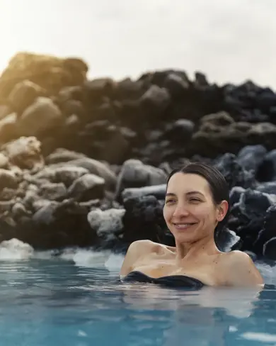 Happy woman enjoying a peaceful soak in the geothermal waters of Blue Lagoon Iceland, surrounded by black lava rocks at sunset—capturing a moment of wellness on a Travel Reykjavik spa experience.