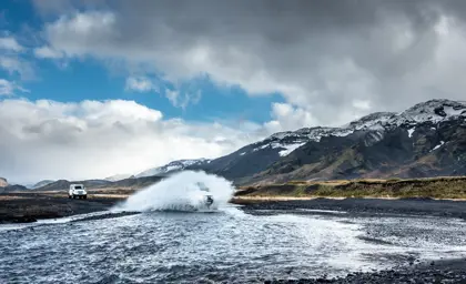 A wide-angle view of the Icelandic highlands as a superjeep from Travel Reykjavík splashes through a river, surrounded by scenic mountains and cloudy skies.